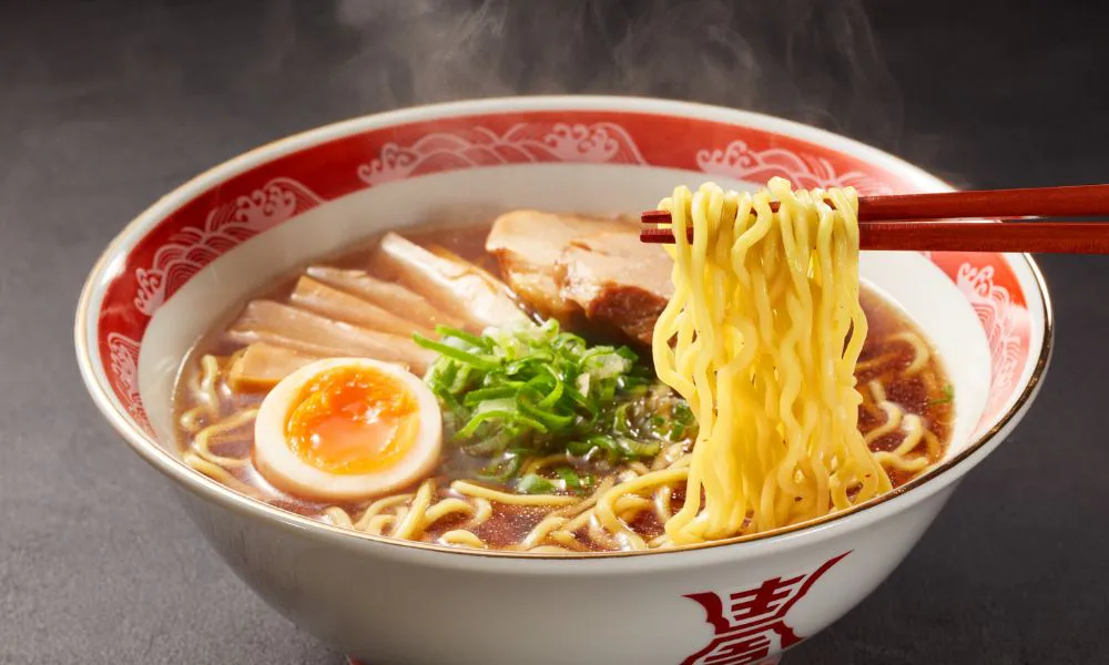 Japanese ramen in red-rimmed bowl, rich broth with soft-boiled egg, chashu and scallions. Red chopsticks lifting noodles, dark background.