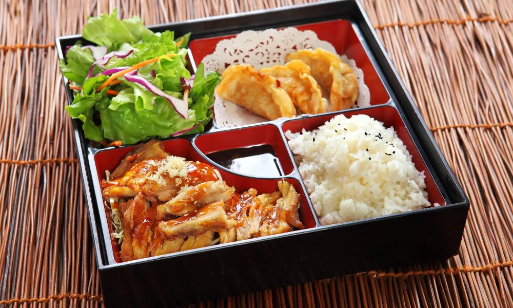 Black divided Japanese bento tray on bamboo mat, filled with glazed teriyaki chicken, sesame-topped steamed rice, fried gyoza dumplings, mixed green salad and small soy sauce compartment.