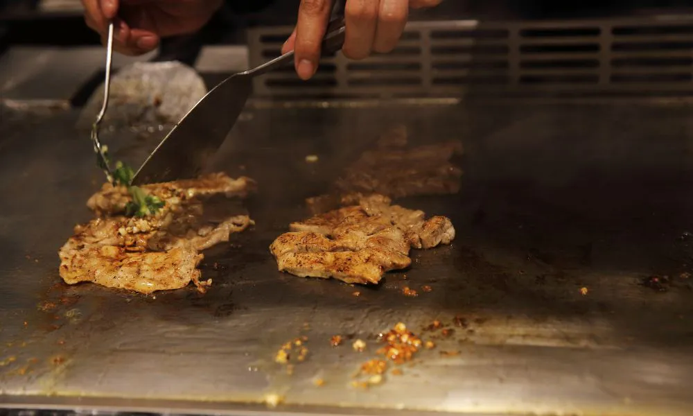 Close-up of chef cooking seasoned meat on teppanyaki grill with metal spatula and tongs, garlic bits on hot metal surface.