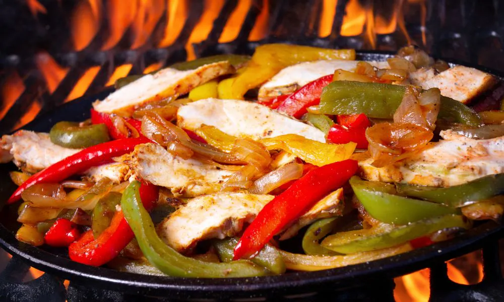 Cast iron sizzling pan filled with seasoned grilled chicken breast strips, mixed red, green and yellow bell peppers plus onions, cooking over bright open grill flames.