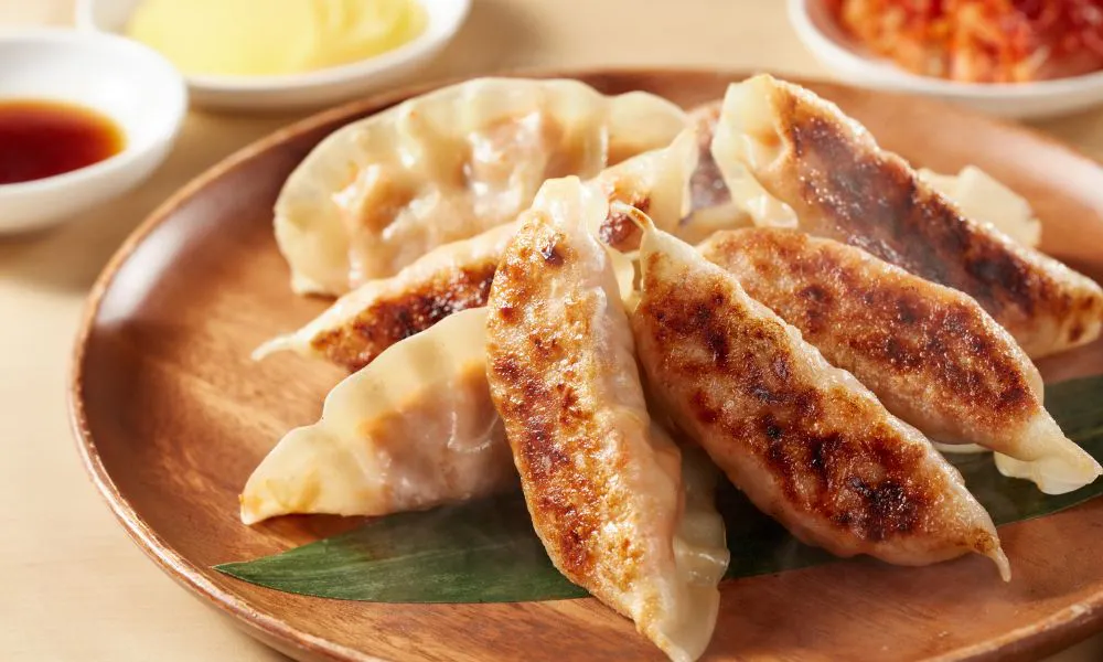Golden crispy pan-fried Japanese gyoza dumplings arranged on wooden plate over green leaf, served with small bowls of soy dipping sauce, kimchi and creamy yellow sauce, faint steam rising.